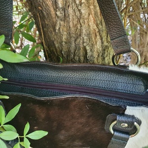 May include: A brown and white cowhide tote bag with a black leather zipper and handles. The bag has a silver ring on each handle.