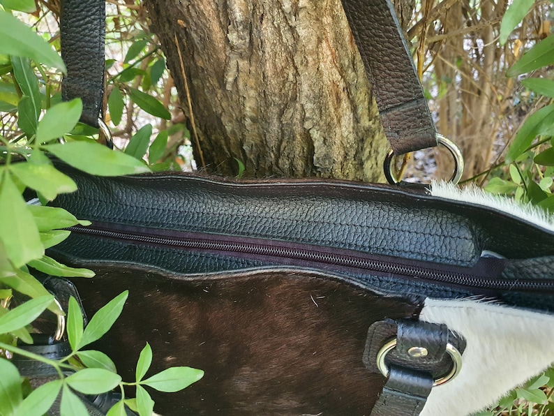 May include: A brown and white cowhide tote bag with a black leather zipper and handles. The bag has a silver ring on each handle.