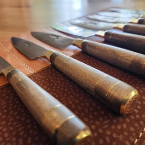 May include: A set of six kitchen knives with wooden handles and gold accents are displayed in a brown leather roll. The knives are arranged in order of size, with the largest knife at the top and the smallest knife at the bottom.