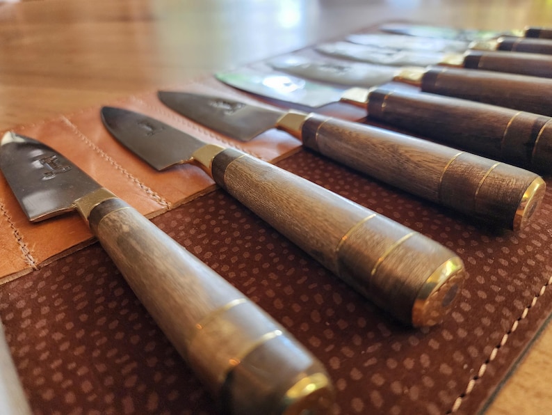 May include: A set of six kitchen knives with wooden handles and gold accents are displayed in a brown leather roll. The knives are arranged in order of size, with the largest knife at the top and the smallest knife at the bottom.
