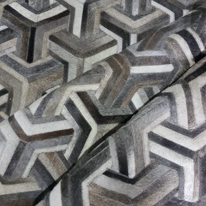May include: A geometric patterned rug made from different shades of grey, brown, and white cowhide. The rug is made up of a series of interlocking triangles and squares.