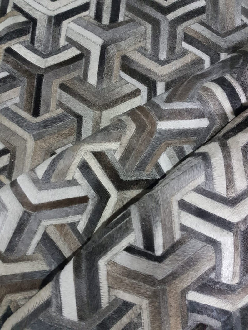 May include: A geometric patterned rug made from different shades of grey, brown, and white cowhide. The rug is made up of a series of interlocking triangles and squares.