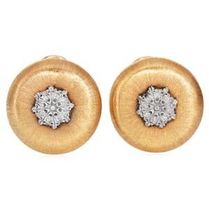 May include: A pair of gold-tone stud earrings with a textured, circular design. Each earring features a silver-colored, snowflake-like center. The earrings are set against a white background.