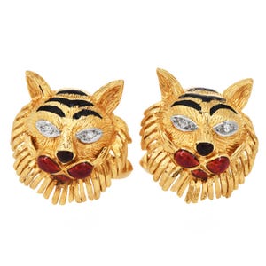 May include: A pair of gold-toned tiger head earrings. The earrings feature black stripes, red accents, and diamond-like embellishments for eyes. The earrings have a textured, detailed design.