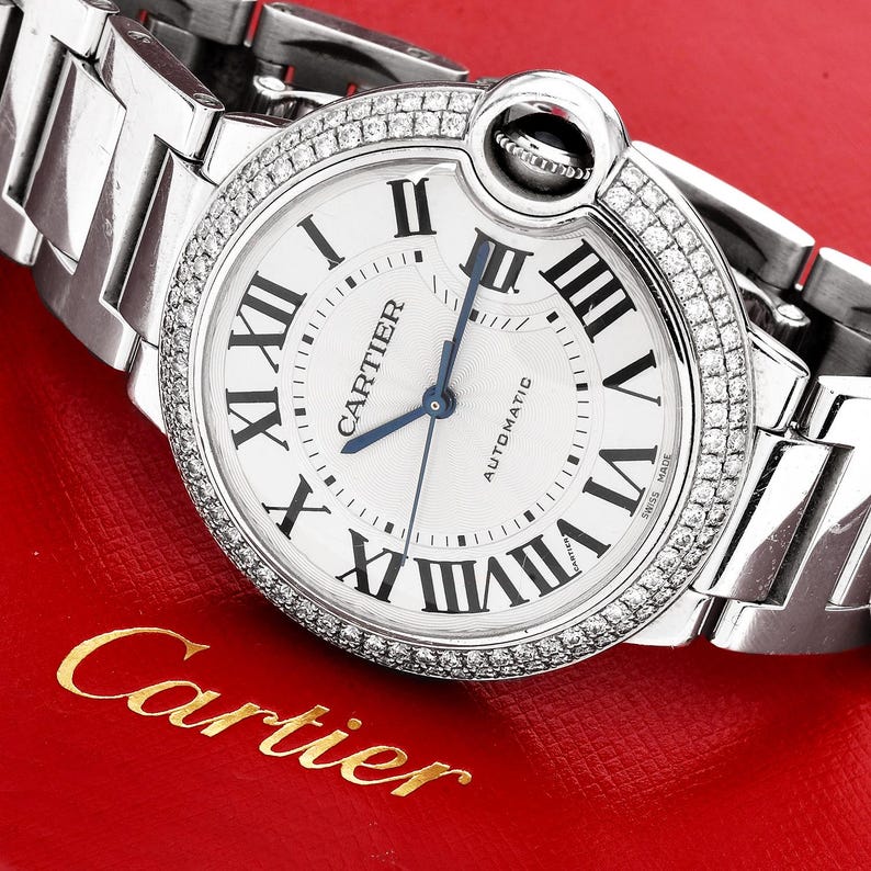 Puede incluir: Un reloj Cartier plateado con una esfera blanca y un bisel de diamantes. El reloj tiene marcadores de hora en n&uacute;meros romanos y una aguja de segundos azul. El reloj es autom&aacute;tico y dice "Swiss Made" en la esfera.