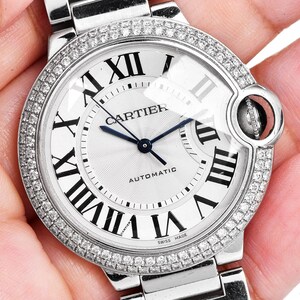 Puede incluir: Un reloj Cartier plateado con una esfera blanca y un bisel de diamantes. El reloj tiene marcadores de hora en n&uacute;meros romanos y una ventana de fecha en la posici&oacute;n de las 3 en punto. El reloj es autom&aacute;tico y dice "Swiss Made" en la esfera.