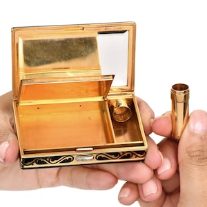 May include: A gold and black compact with a mirror and a small compartment. The compact is open and the mirror is visible. The compact has a black enamel design with gold accents. The compact is labeled 'Emarès New York'.