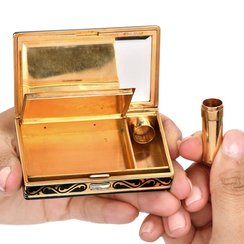 May include: A gold and black compact with a mirror and a small compartment. The compact is open and the mirror is visible. The compact has a black enamel design with gold accents. The compact is labeled 'Emarès New York'.