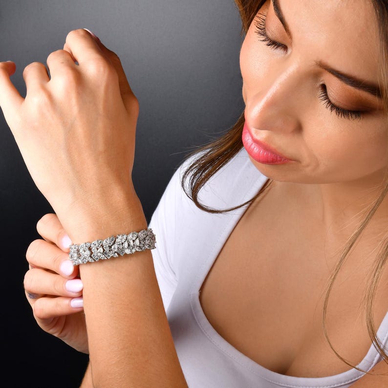 May include: A silver bracelet with a floral design. The bracelet is made of multiple small, sparkling stones. The bracelet is worn on a wrist, and the person is wearing a white top.