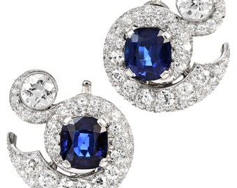 May include: A pair of earrings featuring a blue sapphire gemstone surrounded by a halo of diamonds. Each earring has a smaller diamond set in a swirl design.