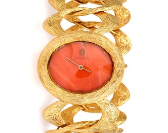 May include: A gold-toned watch with a unique, textured, and linked bracelet. The watch face is round and features a coral-colored dial with the word "Omega" printed on it.