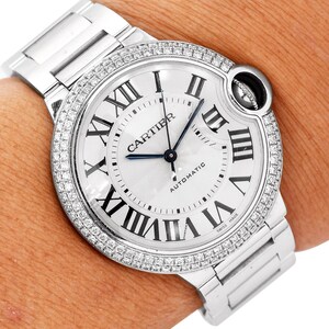 Puede incluir: Un reloj Cartier plateado con un bisel engastado con diamantes. La esfera del reloj es blanca con marcadores de hora de n&uacute;meros romanos y una aguja de segundos azul. El reloj tiene una ventana de fecha en la posici&oacute;n de las 3 en punto y las palabras "Cartier" y "Automatic" impresas en la esfera. El reloj est&aacute; en una pulsera plateada.