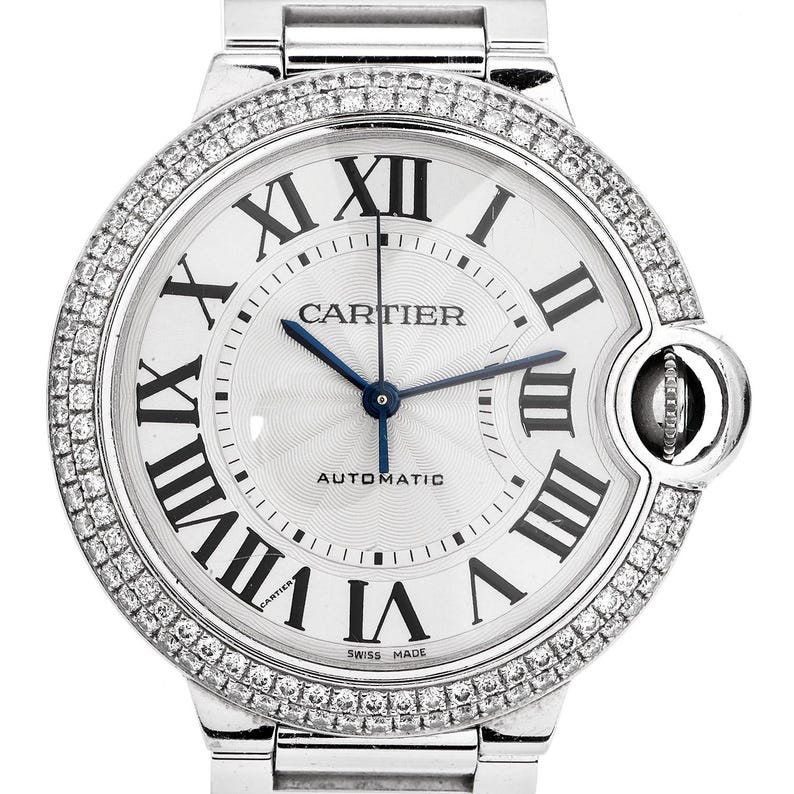 Puede incluir: Un reloj Cartier Ballon Bleu plateado con un bisel engastado con diamantes y una esfera blanca. El reloj presenta marcadores de hora en n&uacute;meros romanos, una aguja de acero azul y las palabras "Cartier" y "Automatic" impresas en la esfera.
