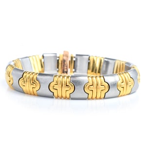 May include: A two-tone gold and silver bracelet with a repeating pattern of interlocking circles. The bracelet is made of metal and has a polished finish.