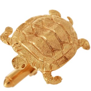 May include: A gold-colored turtle-shaped cufflink. The cufflink features a detailed design of a turtle's shell, head, and flippers. The cufflink is made of a shiny, metallic material and is set against a white background.