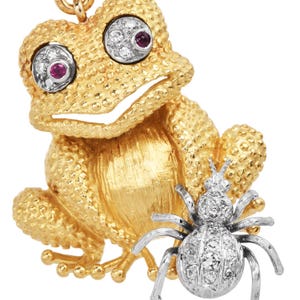 May include: A gold-toned frog pendant with textured skin and ruby eyes, accented with diamond detailing. A silver-toned spider charm is attached, also adorned with diamonds. The pendant is on a white background.