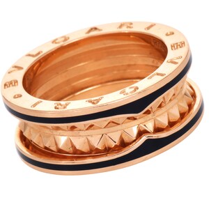 May include: A gold-toned ring with black enamel accents. The ring features a multi-layered design with engraved lettering and a geometric pattern. The ring is a luxury jewelry item.
