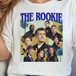 Puede incluir: Camiseta blanca con un gráfico de la serie de televisión "The Rookie". El diseño incluye el título del programa en azul y un collage de actores sobre un fondo amarillo y azul. Una prenda casual para el día a día.