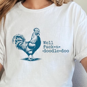 Może przedstawiać: Biała koszulka z niebieską grafiką koguta i napisem "Well fuck-a-doodle-doo". Koszulka wykonana jest z miękkiego materiału i ma krótkie rękawy. Modelka ma na sobie niebieskie dżinsy.