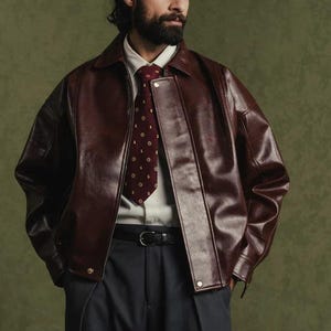Può includere: Giacca in pelle bordeaux con colletto e dettagli con bottoni. La giacca è indossata sopra una camicia bianca e una cravatta bordeaux fantasia. La persona indossa anche pantaloni blu scuro e una cintura nera.