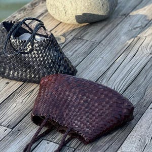 May include: Two woven tote bags, one in black and one in dark brown, featuring a consistent basket-weave pattern throughout the body and matching braided handles. The bags have an open top design and a structured, rectangular base shape.