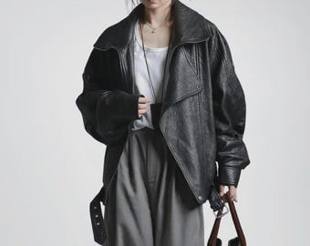 May include: A black leather jacket with a wide collar and a belt at the waist. The jacket has a textured appearance and a slightly oversized fit. The model is wearing a white tank top and gray pants. A brown handbag is held in the hand.