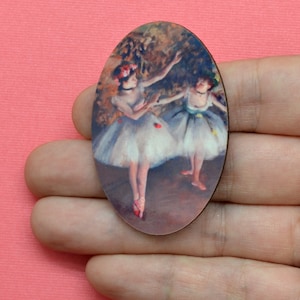 May include: Oval-shaped wooden pendant featuring a painting of two ballerinas in white tutus and pink pointe shoes. The artwork has a soft, impressionistic style with a muted color palette. The ballerinas are depicted in a dance pose.