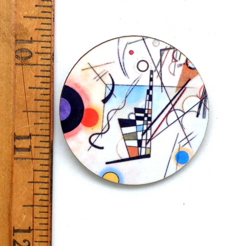 A round pin featuring the artwork of Wassily Kandinsky Composition. It has a white background with lines, circles, squiggles and arcs. The colors are blue, purple, black, red and yellow. It is next to a ruler showing the Diameter is 1-1/2 inches.