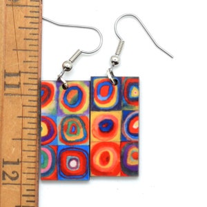 Each earring has squares, that are filled with circles. They are created with imperfect circles in a rainbow of colors including orange, red, blue, yellow, purple and green. The earrings are next to a ruler that show they are 1-1/8 inch tall.