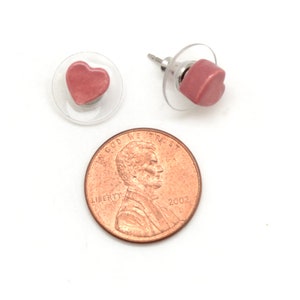 May include: Two pink heart-shaped stud earrings with clear plastic backings. A copper penny is next to the earrings for size comparison. The penny is dated 2002 and has the words "In God We Trust" and "Liberty" on it.