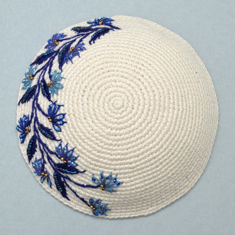 This is a round crochet yarmulke that the base is a off white  natural color.  Around about 45% of the edge, there are embroidered blue leaves with dark and light blue flowers. The leaves and flowers have tiny accent beads of blue and bronze.