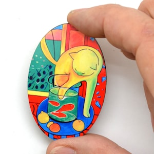 May include: Oval wooden brooch depicting a yellow cat reaching into a glass bowl with red fish. The background shows a window, fruit, and a blue table, with a red and green colour scheme.
