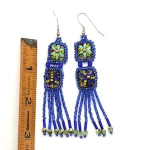 May include: Handmade beaded earrings with a floral design. The earrings feature blue, green, and gold beads, with a square floral design at the top and bottom. The earrings have a fringe of blue beads and measure approximately 7.6 cm in length.