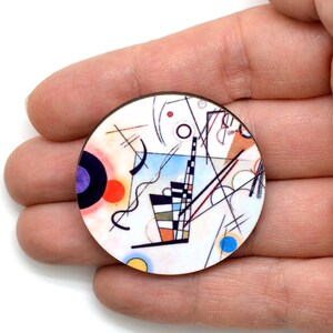 A round pin featuring the artwork of Wassily Kandinsky Composition. It has a white background with lines, circles, squiggles and arcs. The colors are blue, purple, black, red and yellow.  It is resting on fingers.
