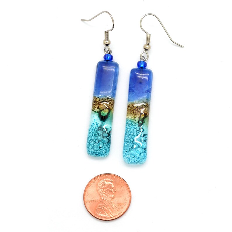 Skinny rectangle glass blue earrings. The earrings are next to a USA copper penny showing the penny is much wider than the earrings and not as tall.