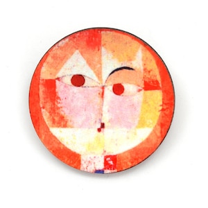May include: A circular brooch featuring an abstract face design with a vibrant red border. The face is composed of geometric shapes in shades of pale pink, yellow, and white, with two prominent red circular eyes and a small dark mark for a mouth.