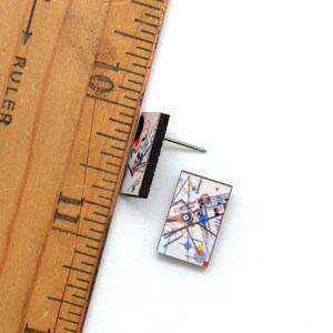 Small rectangle stud earrings featuring Kandinsky geometric art in a rainbow of circles, lines and squiggles. The earrings are next to a ruler showing they are 5/8 inch long. One earring is on the side revealing the dark brown, almost black stain.