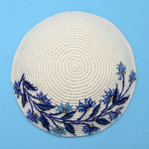 This is a round crochet yarmulke that the base is a off white  natural color.  Around about 45% of the edge, there are embroidered blue leaves with dark and light blue flowers. The leaves and flowers have tiny accent beads of blue and bronze.