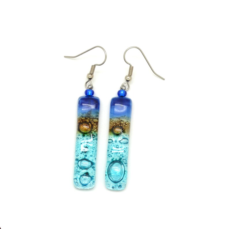 Skinny rectangle glass blue earrings. The very top has a cobalt blue glass bead. The fused glass is in three bands of color. At the top is dark cobalt blue. Second, bronze. Third and widest is an aqua turquoise color. The glass has bubbles.