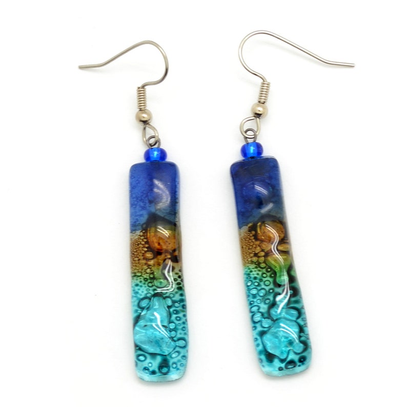 Skinny rectangle glass blue earrings. The very top has a cobalt blue glass bead. The fused glass is in three bands of color. At the top is dark cobalt blue. Second, bronze. Third and widest is an aqua turquoise color. The glass has bubbles.