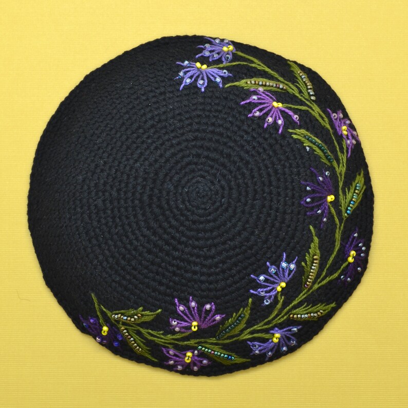 A round black color crochet yarmulke. Around the rim on about 50% of the kippa are embroidered flowers with stems and leaves in assorted shades of purple. There are glass seed bead accents on the leaves and flowers that are purple and yellow.