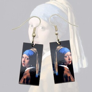 May include: A pair of rectangular earrings featuring the image of the painting "Girl with a Pearl Earring" by Johannes Vermeer. The earrings have a black background and a silver hook.