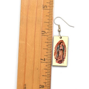 A rectangle earring with an image of the virgin of Guadalupe is next to a ruler showing the drop is about 1-1/8 inch long.