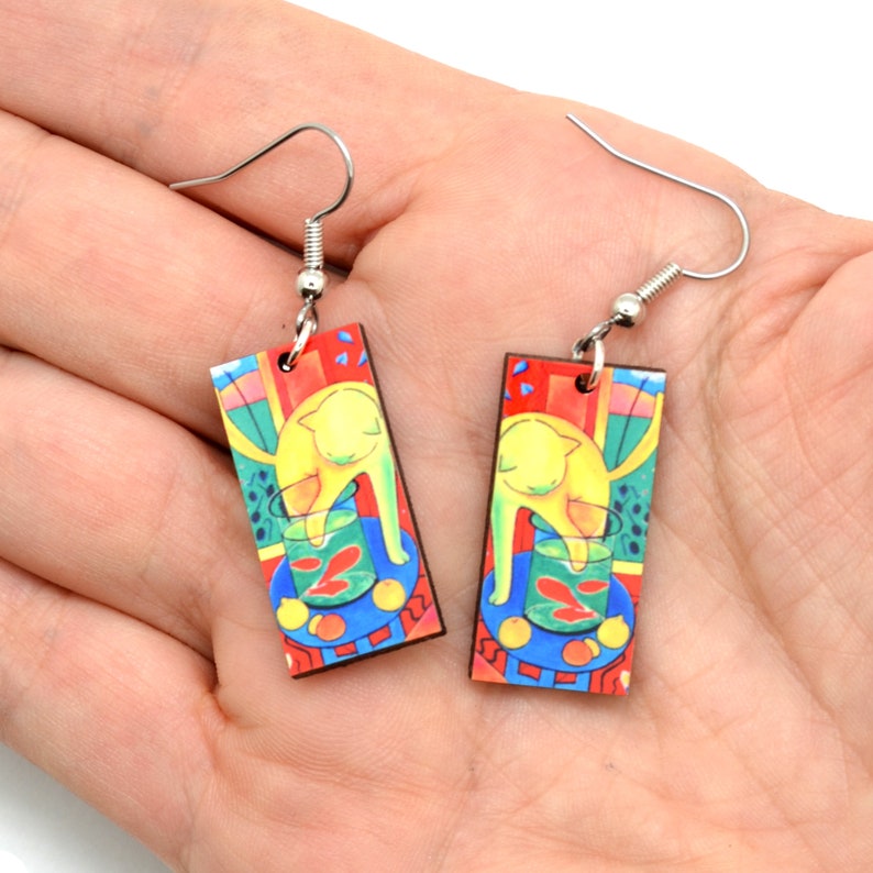 Rectangle earrings featuring the bright artwork of Matisse in the palm of a hand. There is a yellow cat with it's paw in a fish bowl with red fish. The painting has bright green, yellow, red and blue.