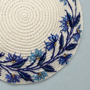 Detail shown of blue embroidered and beaded flowers and leaves that adorn an egg-shell color crochet kippa.