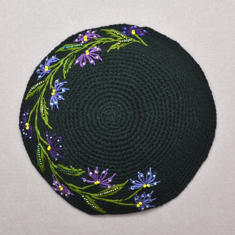 A round black color crochet yarmulke. Around the rim on about 50% of the kippa are embroidered flowers with stems and leaves in assorted shades of purple. There are glass seed bead accents on the leaves and flowers that are purple and yellow.