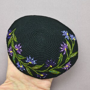 A round black color crochet yarmulke. Around the rim on about 50% of the kippa are embroidered flowers with stems and leaves in assorted shades of purple. The yarmulke is held in a hand.