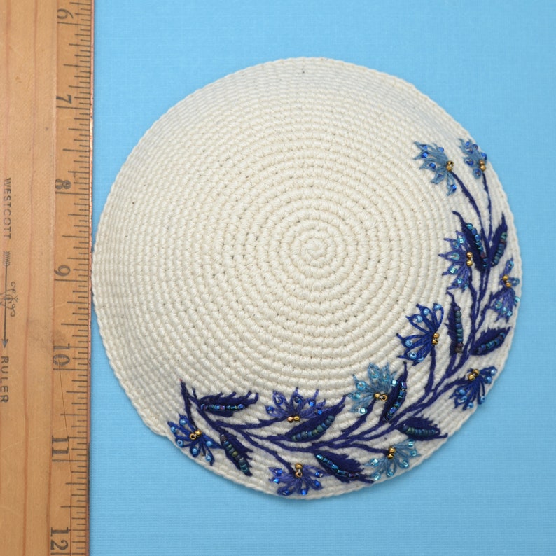 This is a round crochet yarmulke that the base is a off white  natural color.  Around about 45% of the edge, there are embroidered blue leaves with dark and light blue flowers. A ruler shows the kippa is about 5-1/2 inches wide.
