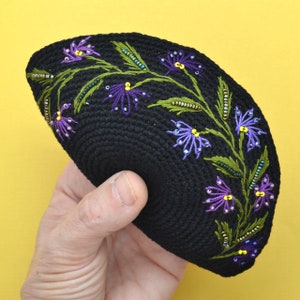 A round black color crochet yarmulke. Around the rim on about 50% of the kippa are embroidered flowers with stems and leaves in assorted shades of purple. The yarmulke is folded and held in a hand so you can see the detail closer up.