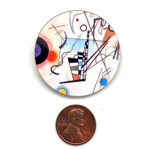 A round pin with the artwork of Kandinsky next to a USA copper penny for size comparison. The pin is much larger than a penny.